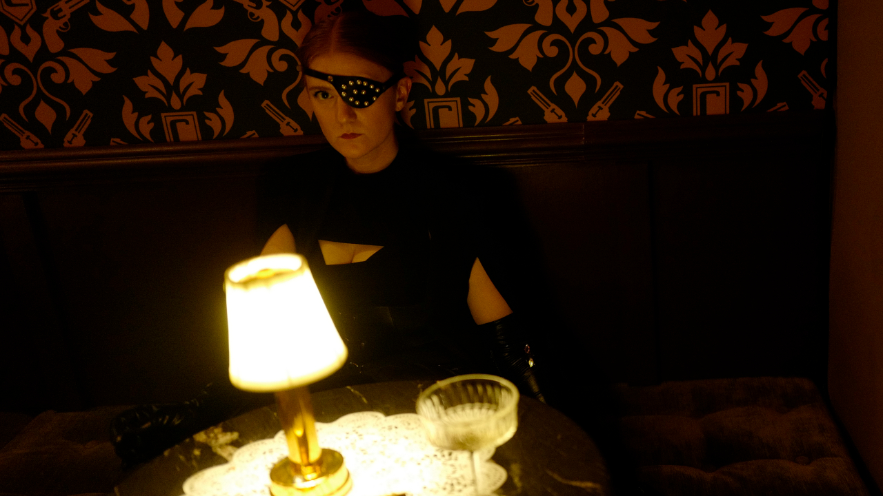 Eine geheimnisvolle Frau mit Augenklappe sitzt an einem schwach beleuchteten Tisch im The Continental.