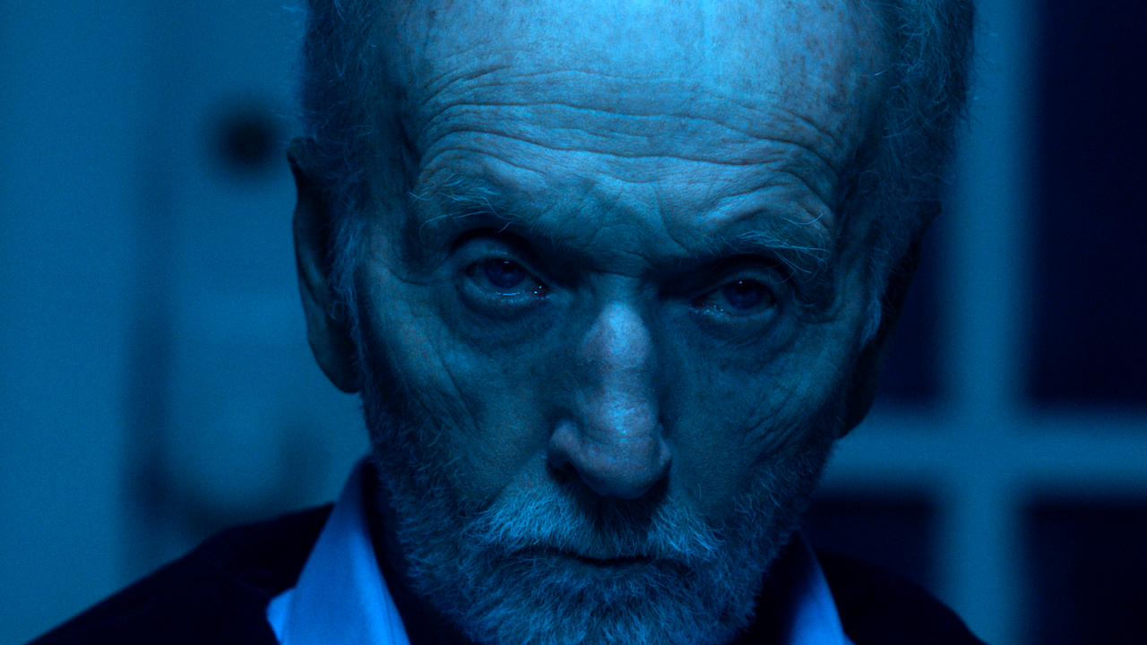 Tobin Bell verzieht das Gesicht, während er in Saw X in einem blau erleuchteten Raum sitzt.