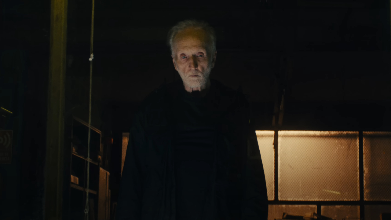 Tobin Bell steht in Saw X bedrohlich in einem Lagerhaus, schwarz gekleidet.