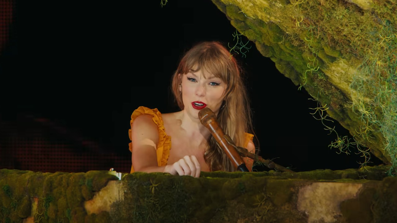 Ein Screenshot von Taylor Swift, wie sie während des Trailers zum Konzertfilm „The Eras Tour“ in ein Mikrofon spricht, während sie hinter einem Klavier sitzt.