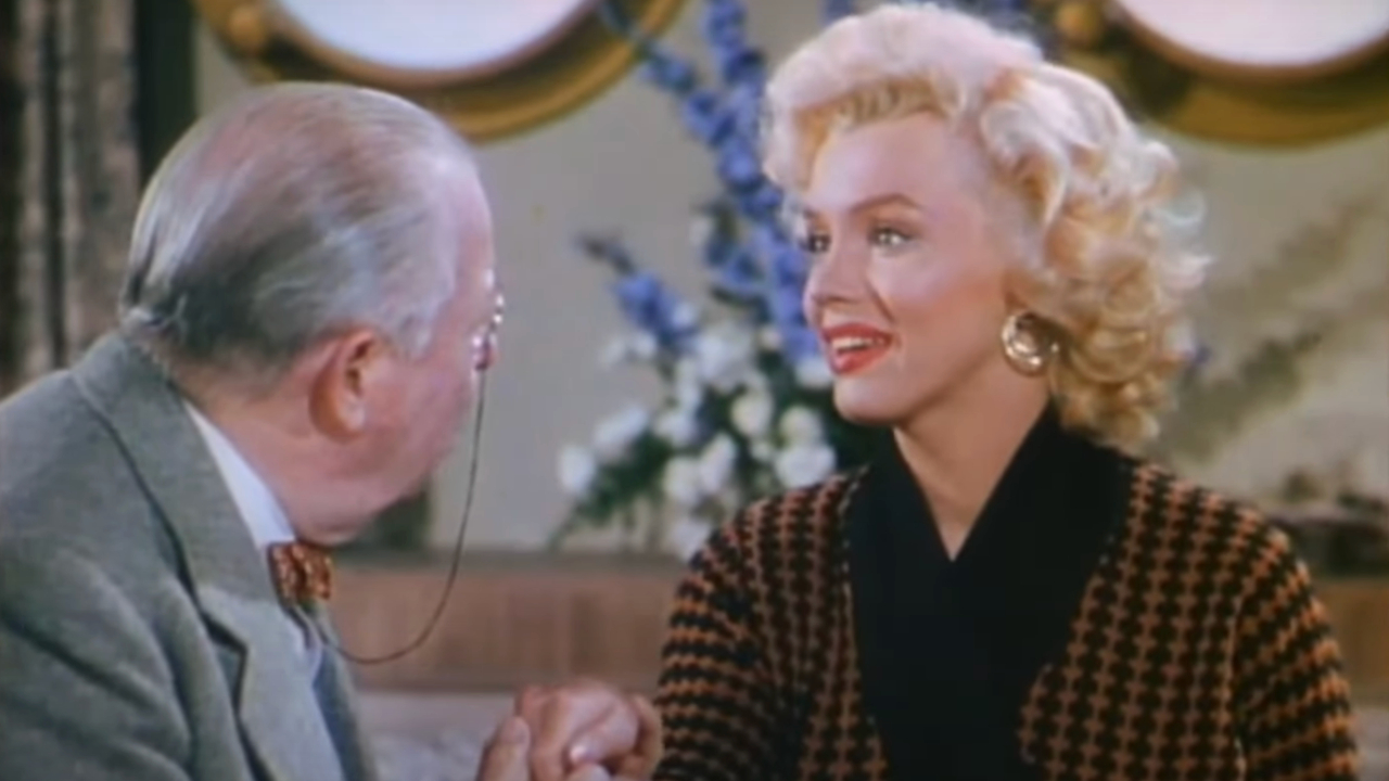 Marilyn Monroe sitzt lächelnd da, während ein alter Mann in Gentlemen Prefer Blondes ihre Hand hält.