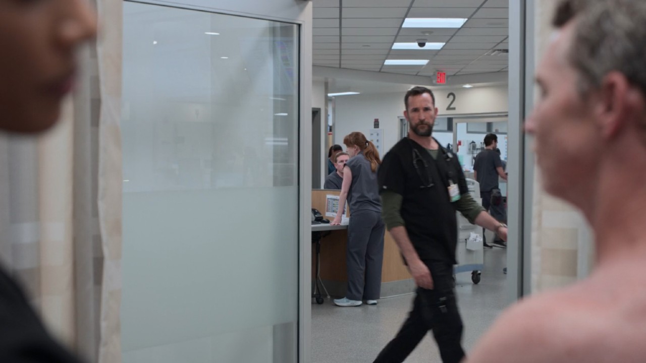 Le Dr Robby passe devant une porte ouverte pendant que le Dr Mohan aide le Dr Abbot sur The Pitt.