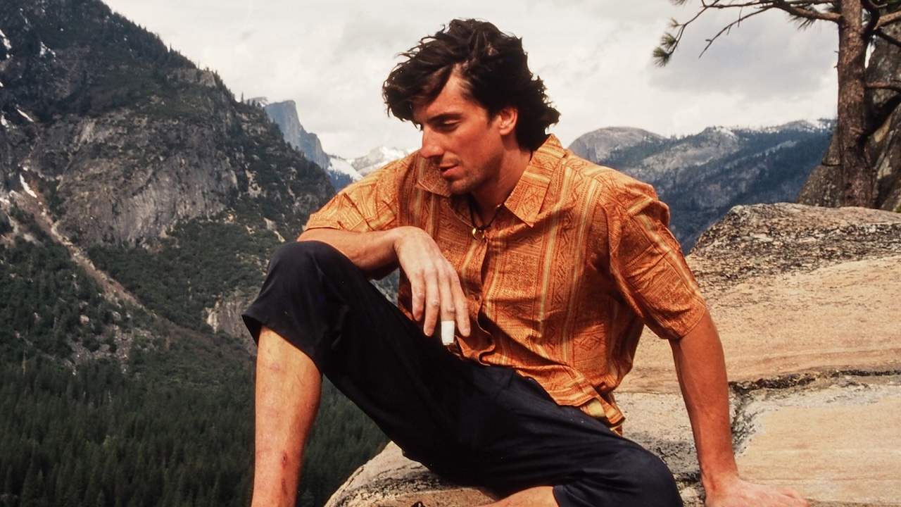 Дин Поттер сидит в оранжевой рубашке и черных брюках на фоне гор.