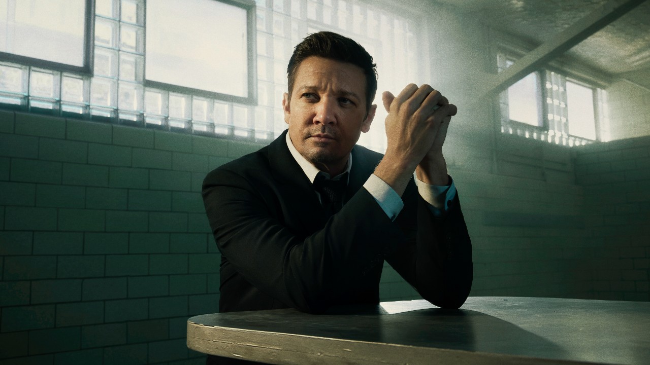 Jeremy Renner sitzt mit gefalteten Händen an einem Tisch, während seine Ellbogen auf dem Tisch ruhen.