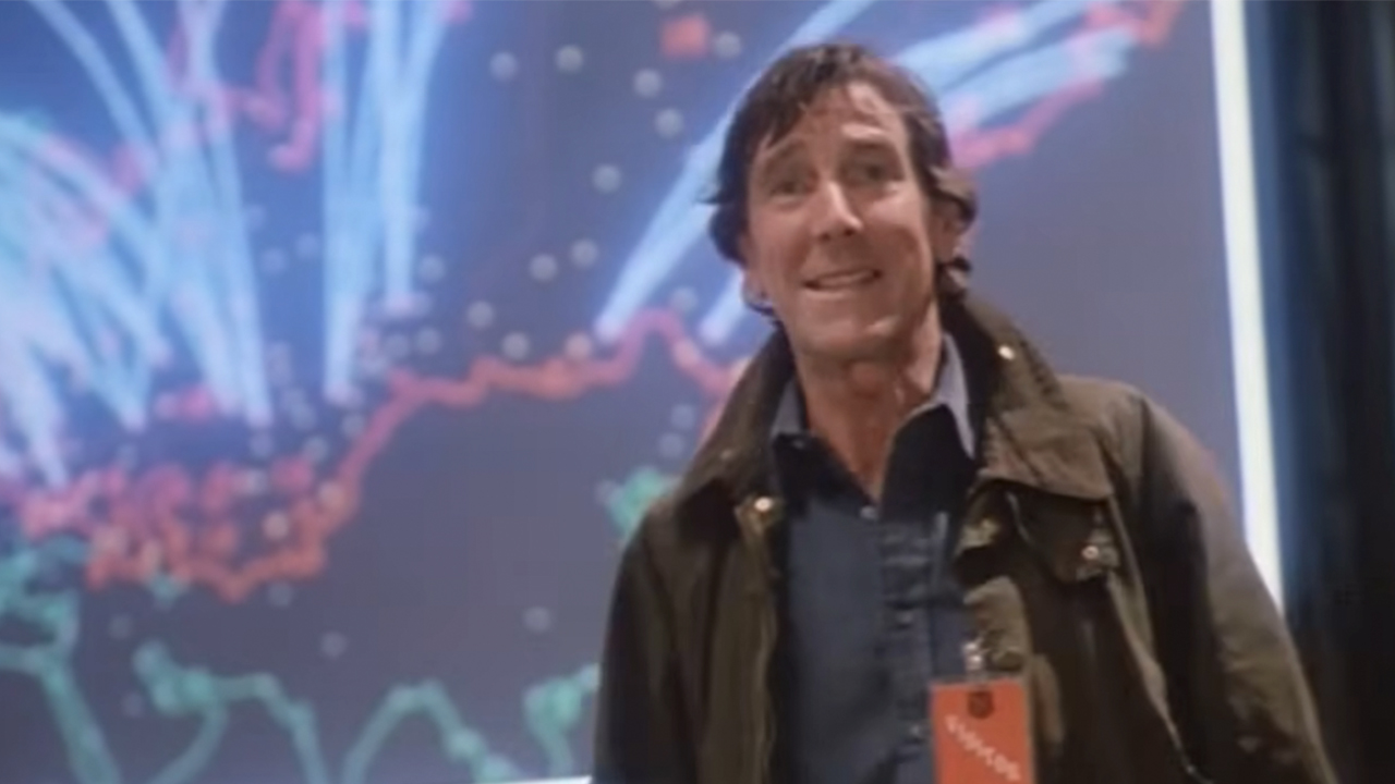 John Wood steht und verzieht das Gesicht vor einem Fernsehbildschirm mit bunten Linien, die Raketenbahnen in WarGames zeigen