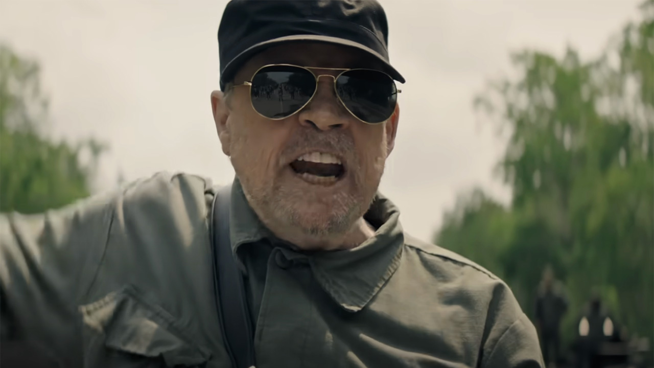 Mark Hamill in Sonnenbrille, Arbeitskleidung und Hut brüllt draußen in „The Long Walk“.